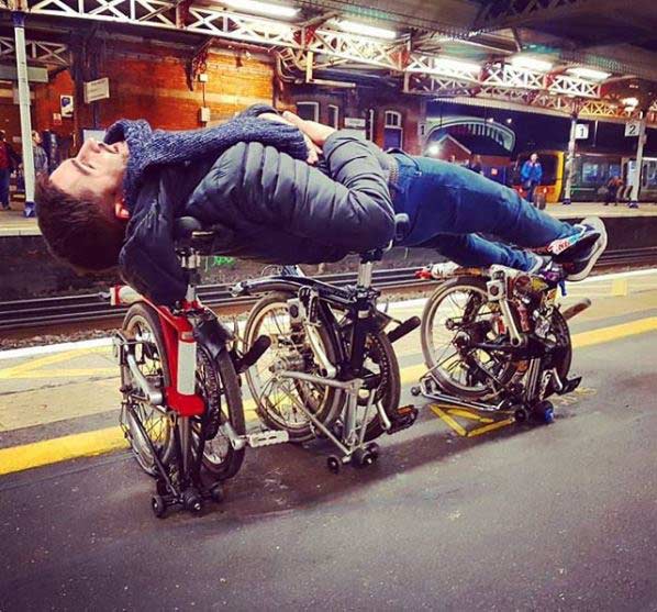 Person lying on top of 3 folded Bromptons on railway platform