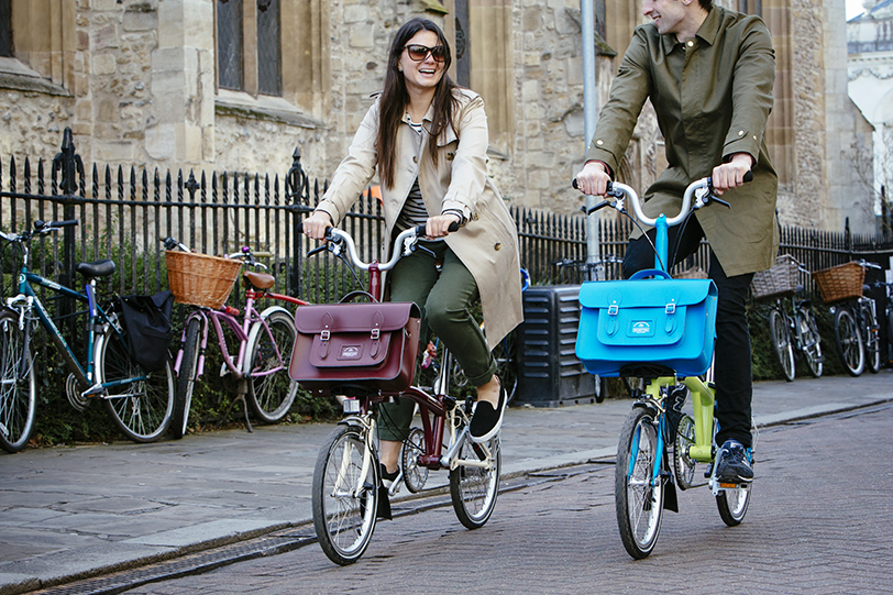 Two people riding Brompton bikes with Brompton x Cambridge Satchel Company satchels attached