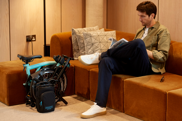 Image of a folded Brompton electric next to man reading on a couch